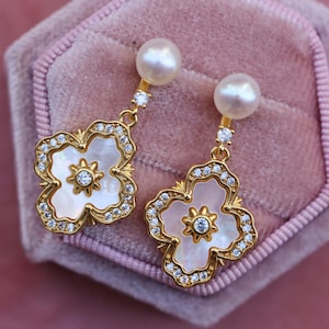 May include: A pair of gold earrings with a white pearl and a white flower-shaped pendant with clear rhinestones.