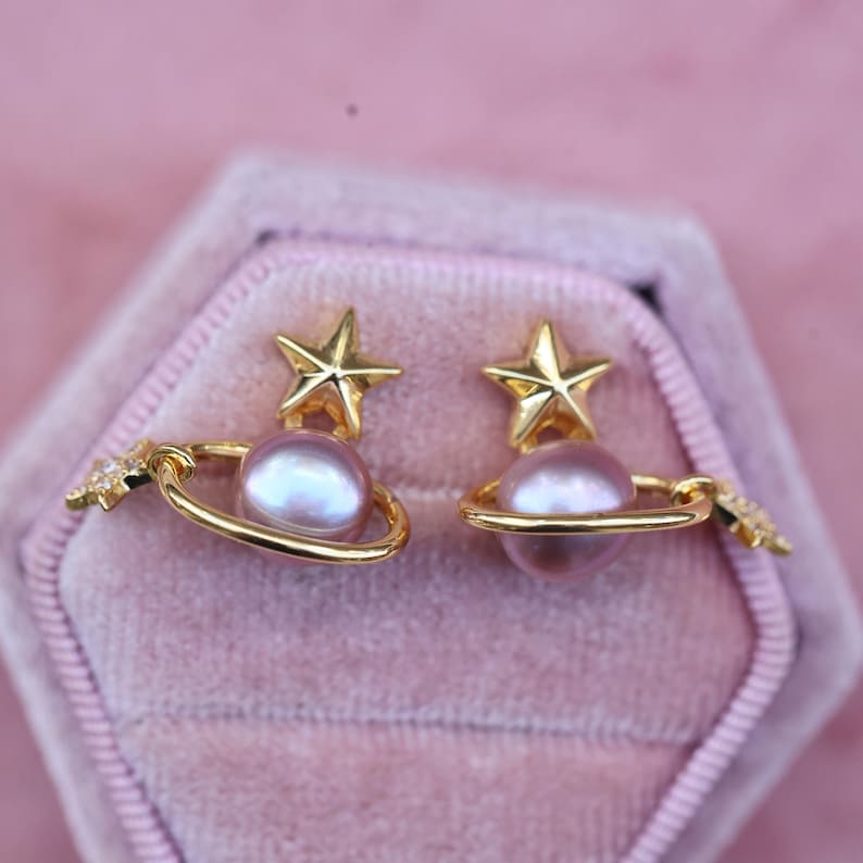 May include: A pair of gold-colored earrings featuring a star and planet design. Each earring has a gold star on top, a light purple pearl, and a gold ring around the pearl. A small star is attached to the ring. The earrings are displayed in a pink velvet box.