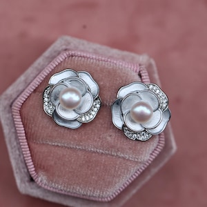 May include: A pair of flower-shaped stud earrings. Each earring features a central, light pink pearl surrounded by white petals and a ring of small, clear crystals. The earrings are set against a pink velvet background.