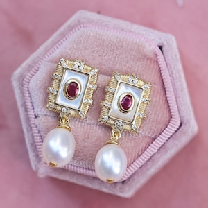 May include: A pair of gold-toned earrings with a rectangular frame design. Each earring features a white center with a red gemstone, surrounded by small clear stones. A large, white pearl dangles from each earring, set against a pink velvet background.
