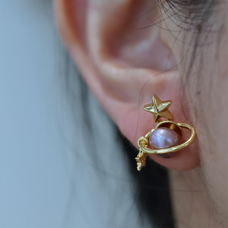 May include: Gold-colored stud earrings featuring a planet design with a pink pearl and a star. The earrings have a celestial theme, with a small star charm dangling below the planet. The earrings are worn on an ear.
