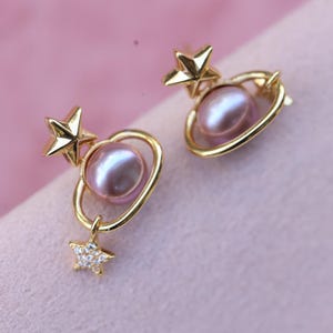 May include: A pair of gold-colored earrings featuring a star and planet design. Each earring has a gold star, a pink pearl, and a gold ring around the pearl. A smaller star with clear stones hangs below the planet.
