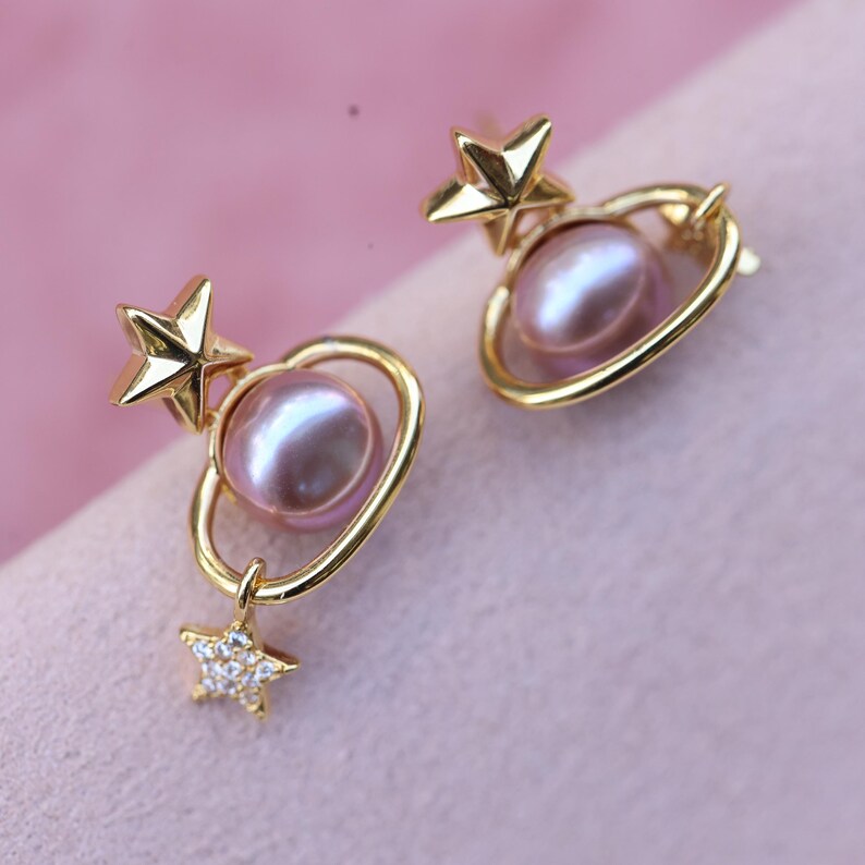 May include: A pair of gold-colored earrings featuring a star and planet design. Each earring has a gold star, a pink pearl, and a gold ring around the pearl. A smaller star with clear stones hangs below the planet.