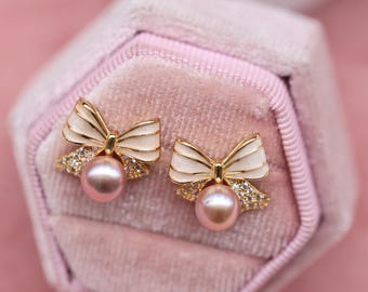May include: A pair of gold-toned bow earrings with white enamel detailing and a central pink pearl. The earrings are set with small, sparkling crystals and are displayed in a pink velvet jewelry box.