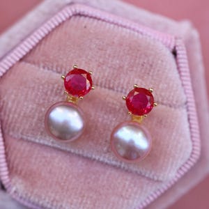 May include: A pair of earrings with a red gemstone and a pearl drop. The earrings are gold-toned and have a simple design.