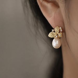 May include: A gold-coloured earring with a leaf and flower design, adorned with small, clear gemstones. A large, white pearl dangles beneath the floral elements. The earring is worn on an ear.