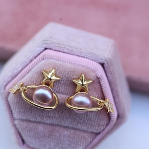 May include: A pair of gold-colored earrings featuring a planet design with a star on top and a small star with crystals on the side. The earrings have a pink pearl in the center and are displayed in a pink velvet jewelry box.