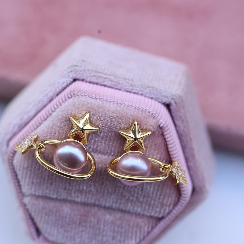 May include: A pair of gold-colored earrings featuring a planet design with a star on top and a small star with crystals on the side. The earrings have a pink pearl in the center and are displayed in a pink velvet jewelry box.