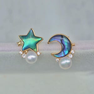 May include: A pair of gold earrings with a star and crescent moon design. The star and moon are made of iridescent abalone shell and are accented with small clear crystals. Each earring has a single white pearl dangling from the bottom.