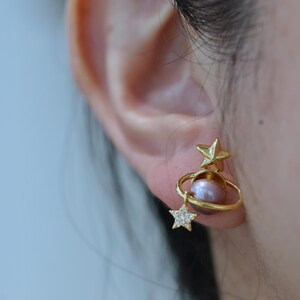 May include: Gold-toned earrings featuring a celestial design. Each earring has a star at the top, a ring encircling a pink pearl, and a dangling star with small, clear stones. The earrings are worn on an ear.
