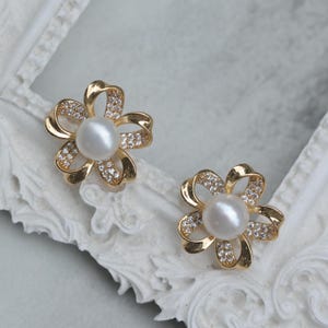 以下が含まれることがあります： 花の形をした金のイヤリングのペア。それぞれの金のフラワーモチーフの中央に真珠が飾られています。花は小さなクリアなラインストーンで飾られています。