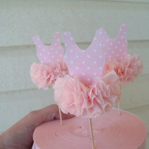 May include: Pink ballerina-themed cupcake toppers. Each topper features a pink paper tutu and a pink leotard with white polka dots. The toppers are attached to wooden sticks, perfect for decorating cupcakes or other treats.