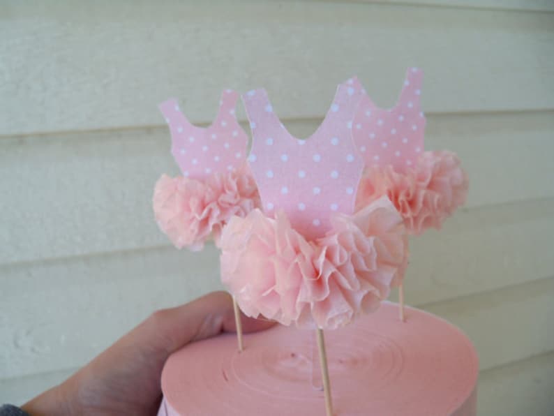May include: Pink ballerina-themed cupcake toppers. Each topper features a pink paper tutu and a pink leotard with white polka dots. The toppers are attached to wooden sticks, perfect for decorating cupcakes or other treats.