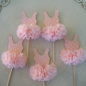 May include: Five pink ballerina tutu cupcake toppers. Each topper features a light pink leotard with white polka dots and a ruffled tutu. The toppers are on wooden sticks, perfect for decorating cupcakes or other treats.