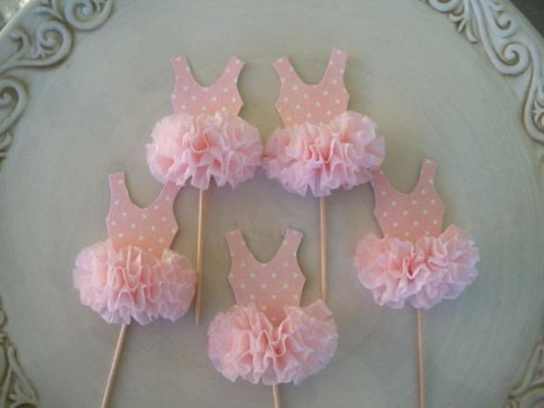 May include: Five pink ballerina tutu cupcake toppers. Each topper features a light pink leotard with white polka dots and a ruffled tutu. The toppers are on wooden sticks, perfect for decorating cupcakes or other treats.