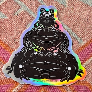 Puede incluir: Una pegatina holográfica con una pila de tres ranas negras. La pegatina tiene un borde iridiscente arcoíris y el texto "@Standing Beetle Art". Las ranas están estilizadas con detalles blancos.