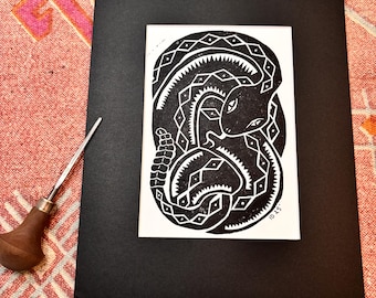 May include: Black and white linocut print of a coiled rattlesnake with a diamond pattern on its body. The print is matted in black and has the text "10.25" in the bottom right corner.