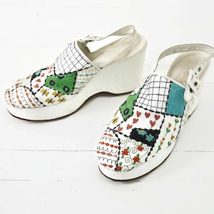 Puede incluir: Un par de zuecos de plataforma blancos con un diseño de patchwork que presenta coloridos patrones florales y geométricos. Los zuecos tienen una suela de plataforma blanca gruesa y una puntera cerrada.