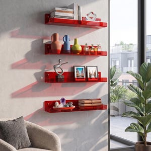 May include: Red acrylic floating shelves mounted on a white wall, displaying books, vases, framed photos, and decorative objects. The shelves are arranged in a vertical line, creating a modern and minimalist aesthetic. A comfortable armchair and a potted plant are in the foreground.