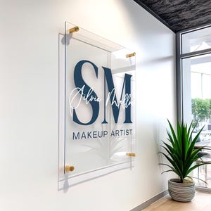 May include: Clear acrylic sign with gold standoffs, featuring navy blue initials "SM", white script name "Silvia Mella", and navy blue text "MAKEUP ARTIST". The sign is mounted on a white wall, suitable for a beauty salon.