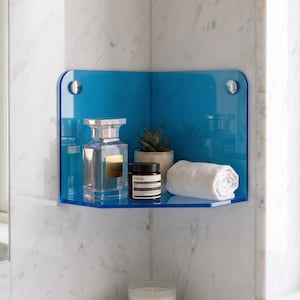 Pode incluir: Prateleira de canto de casa de banho moderna em acrílico azul transparente. Contém um frasco de perfume, uma pequena planta em vaso, um frasco e uma toalha branca enrolada. A prateleira é montada numa parede de mármore.