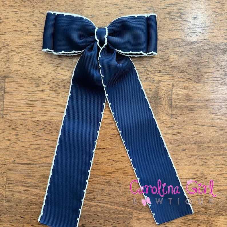 May include: Navy blue bow with white stitching and long tails. The bow is tied in a classic style and has a white trim around the edges. The bow is made of a satin-like material and has a slight sheen.