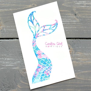 May include: A white card features a colorful mermaid tail design. The tail is a mix of blue, pink, and green patterns. The text "Carolina Girl BWTiQUE" is printed in pink on the card.