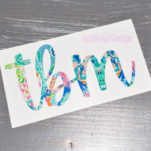 May include: White sticker with the letters "tbm" in a colorful floral pattern. The text "Carolina Girl Boutique" is above the letters in a light pink color. The sticker is on a gray wooden surface.