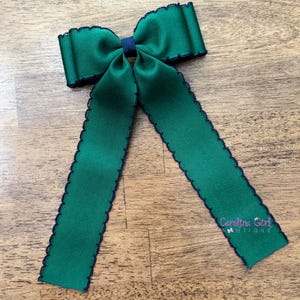 May include: A green ribbon bow with dark blue trim and a dark blue center knot. The ribbon has a scalloped edge and two long, dangling tails. The text "Carolina Girl Boutique" is visible on one of the tails.
