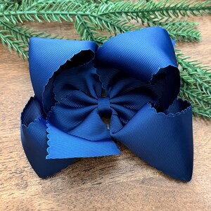 May include: A large, navy blue hair bow with a scalloped edge. The bow is made of a ribbed fabric and has a smaller bow in the center. The bow is likely for a child or a person who likes to wear bows in their hair.