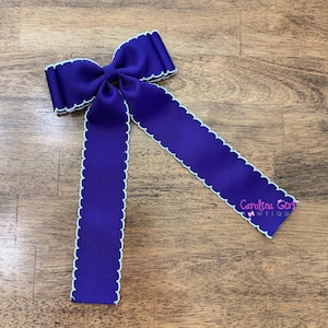 May include: A large, deep purple hair bow with a white scalloped edge. The bow has long, flowing tails and the text "Carolina Girl Wtiqu" printed on one tail. The bow is made of a thick, textured ribbon.