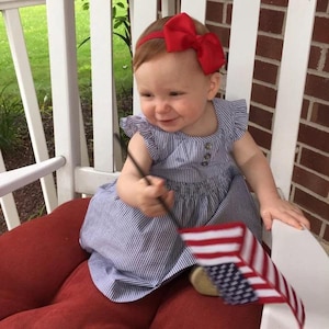 Pu&ograve; includere: Una bambina indossa un vestito a righe blu e bianche con un cerchietto rosso con fiocco e tiene una piccola bandiera americana.