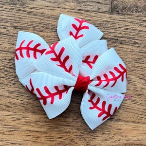 May include: A white hair bow with a baseball design. The bow features a white background with red stitching, and a red center. The bow is made of ribbon and is suitable for a child.