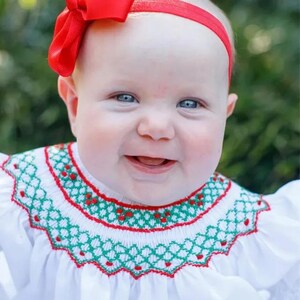 Pu&ograve; includere: Una bambina che indossa un vestito bianco con un colletto ricamato verde e rosso. Indossa una fascia per capelli rossa con un fiocco e ha gli occhi azzurri.