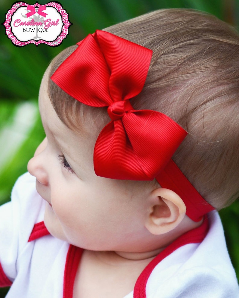 Pu&ograve; includere: Un bambino che indossa una camicia bianca con bordini rossi indossa una fascia per capelli rossa con fiocco. La fascia &egrave; realizzata con un nastro di raso ed &egrave; legata a fiocco sulla parte superiore della testa del bambino.