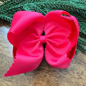 Può includere: Un grande fiocco per capelli rosa acceso con un bordo smerlato. Il fiocco è realizzato in tessuto a coste ed è centrato con una fascia più piccola dello stesso materiale. Aghi di pino verdi sono visibili sullo sfondo.