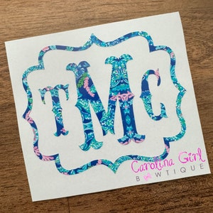 May include: A white sticker featuring the letters "TMC" in a decorative font, filled with a blue, teal, and pink floral pattern. The letters are framed by a scalloped border in a matching pattern. The text "Carolina Girl Bwtique" is printed in pink.