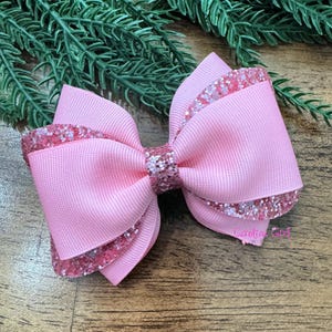 Può includere: Un fiocco per capelli rosa a strati con dettagli glitterati. Il fiocco è realizzato in nastro grosgrain e presenta un fiocco più piccolo al centro. Un accessorio per capelli.