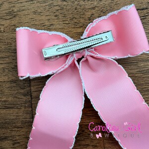 May include: Pink satin bow with white scalloped edges and a silver alligator clip. The bow is tied in a simple knot and has a delicate, feminine look.