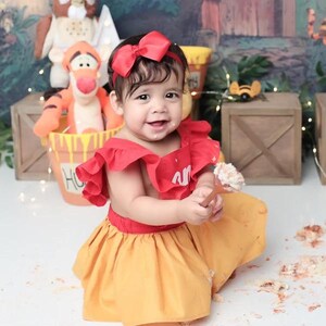 Pu&ograve; includere: Una bambina che indossa un vestito rosso e giallo con un fiocco rosso nei capelli, tiene un cucchiaio con la glassa sopra. Il vestito ha un collo a volant e una gonna gialla. La bambina &egrave; seduta su una superficie bianca con la glassa sopra.