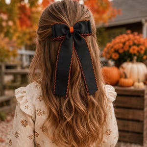 May include: A black hair bow with a vibrant orange scalloped edge and a small orange center knot. The bow features two long, trailing ribbon tails that hang down, providing a decorative accent for hair styled in a half-up, half-down arrangement.