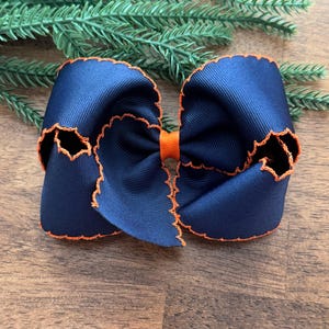 May include: A navy blue hair bow made of grosgrain ribbon with a bright orange stitched scalloped edge and a solid orange center knot. The bow features a layered design with multiple loops, creating a full and voluminous appearance for hair styling.