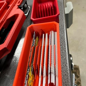 May include: A red plastic organizer with multiple compartments for storing saw blades. The organizer is filled with a variety of saw blades, including yellow, red, and silver blades.