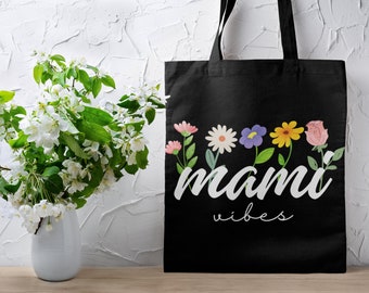 May include: Black tote bag featuring a floral design and the words "mami vibes" in white script. The flowers include pink, white, purple, yellow, and pink blooms. A white vase with white flowers is on the left.