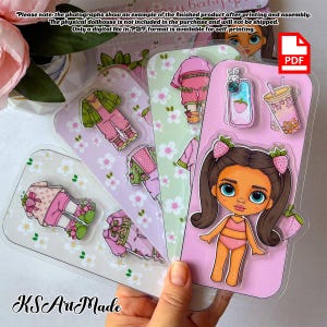 May include: A digital file in PDF format featuring a printable paper doll set. The set includes a doll with brown hair in pigtails, various outfits with strawberry motifs, a smartphone case, and a drink cup, all displayed on floral patterned sheets.