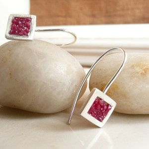 May include: Silver earrings with a square design. Each earring has a square frame filled with small, deep red gemstones. The earrings have a simple hook design for pierced ears, showcasing a modern and minimalist aesthetic.