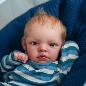 Może przedstawiać: Realistyczna lalka dziecka o jasnej skórze i jasnobrązowych włosach, ubrana w niebiesko-białą koszulkę w paski. Lalka leży w niebieskiej, teksturowanej tkaninie, z skupionym wyrazem twarzy.