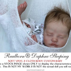Może przedstawiać: Realistyczna lalka dla dziecka z zamkniętymi oczami, otulona białą tkaniną o otwartym splocie. Lalka ma jasnobrązowe włosy i ma na sobie biały strój. Tekst na zdjęciu brzmi "Realborn Daphne Sleeping" i "SOFT VINYL & CLOTH BODY CUSTOM BABY."