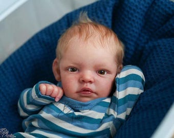 Puede incluir: Una muñeca reborn realista de piel clara y cabello rubio fino, que viste una camiseta de manga larga con rayas horizontales en tonos azul, verde azulado y blanco. La muñeca tiene ojos marrones y descansa sobre una tela azul oscuro texturizada.