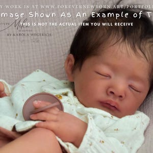 Może przedstawiać: Prototyp lalki reborn, Mika, z zamkniętymi oczami i ciemnymi włosami, ubrana w jasny pajacyk. Obraz zawiera tekst wskazujący, że jest to przykład. Lalka jest autorstwa Karoli Wegerich.